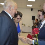 Nie wiem, kto jest na zdjęciu. Przedstawia spotkanie formalne. Osoby wymieniają podarunki, jedna trzyma czerwoną różę.