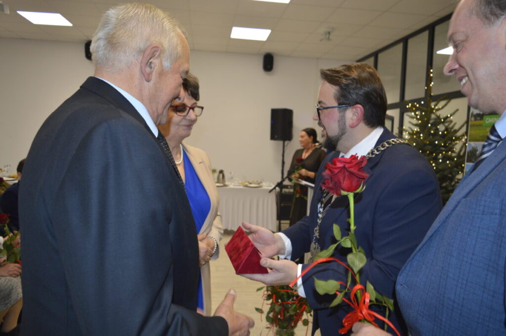 Na zdjęciu widać grupę ludzi podczas uroczystości. Mężczyzna w garniturze wręcza czerwoną różę i czerwone pudełko. W tle choinka i stoły z jedzeniem.