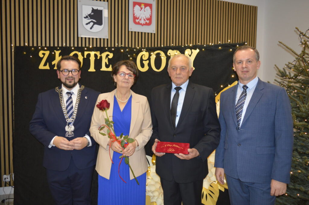 Grupa czterech osób w eleganckich strojach stoi przed czarnym tłem z napisem Złote Gody. Kobieta trzyma czerwoną różę. W tle choinka i złote dekoracje.