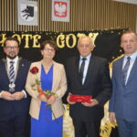 Grupa czterech osób w eleganckich strojach stoi przed czarnym tłem z napisem Złote Gody. Kobieta trzyma czerwoną różę. W tle choinka i złote dekoracje.