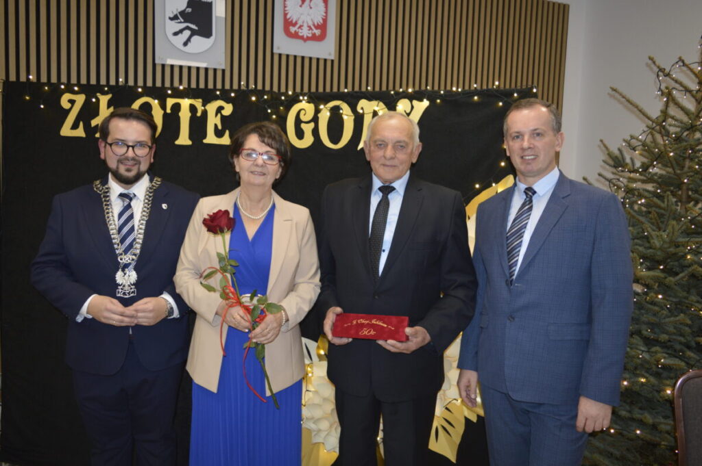 Na zdjęciu widoczne są cztery osoby na tle tablicy z napisem Złote Gody. Osoba trzecia trzyma czerwony certyfikat, a czwarta różę. Z prawej strony stoi ozdobiona światełkami choinka.