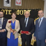Na zdjęciu widoczne są cztery osoby na tle tablicy z napisem Złote Gody. Osoba trzecia trzyma czerwony certyfikat, a czwarta różę. Z prawej strony stoi ozdobiona światełkami choinka.