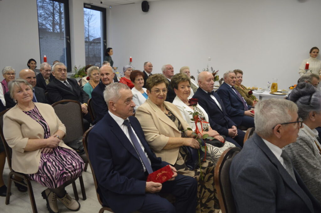 Grupa elegancko ubranych starszych osób siedzi w jasnym pokoju na uroczystości. Pani trzyma czerwoną różę. W tle stół z napojami i ozdobami.