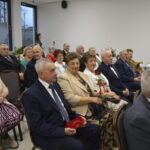 Grupa elegancko ubranych starszych osób siedzi w jasnym pokoju na uroczystości. Pani trzyma czerwoną różę. W tle stół z napojami i ozdobami.