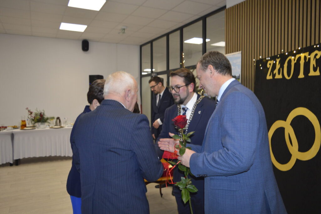Na zdjęciu widać grupę osób w eleganckich garniturach, na pierwszym planie wręczana jest czerwona róża. Tło stanowi biała sala z drewnianymi panelami i stołem z napojami. W prawym rogu widnieje napis ZŁOTE