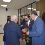 Na zdjęciu widać grupę osób w eleganckich garniturach, na pierwszym planie wręczana jest czerwona róża. Tło stanowi biała sala z drewnianymi panelami i stołem z napojami. W prawym rogu widnieje napis ZŁOTE