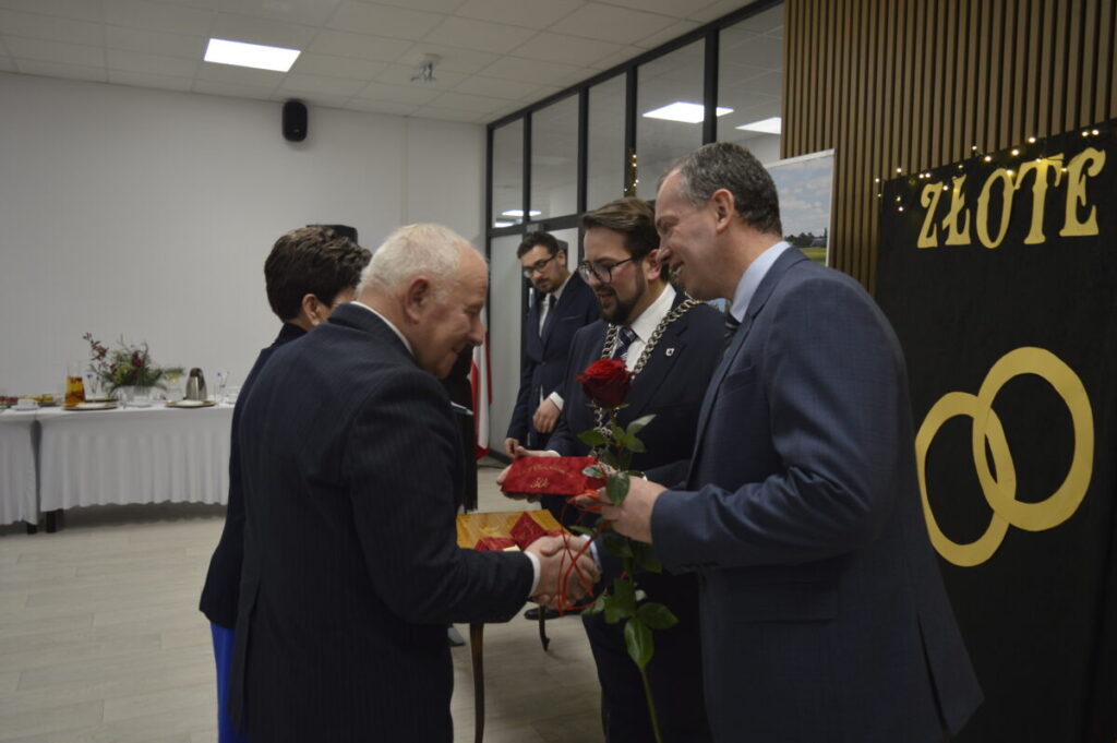 Na obrazie widać ludzi w garniturach, jeden trzyma różę. W tle biały stół z napojami, obok czarna tablica z napisem Złote. Sala ma jasne ściany i sufit.