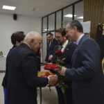 Na obrazie widać ludzi w garniturach, jeden trzyma różę. W tle biały stół z napojami, obok czarna tablica z napisem Złote. Sala ma jasne ściany i sufit.