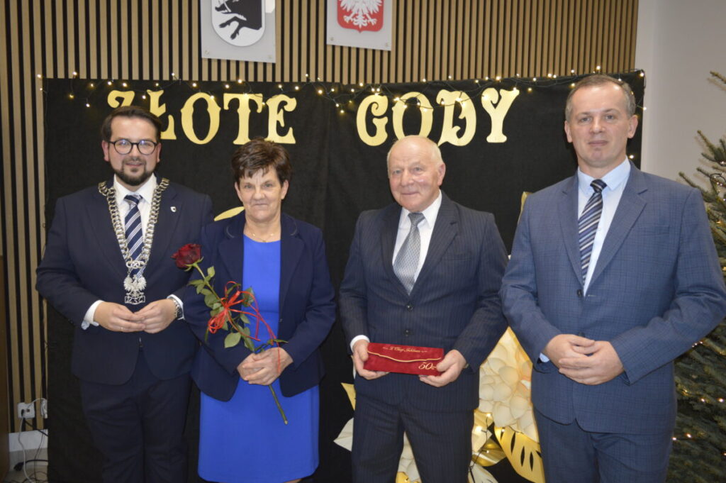 Na zdjęciu są cztery osoby w eleganckich strojach. Tło zdobią napisy Złote Gody oraz świąteczne dekoracje. Dominują kolory ciemne z elementami złota.