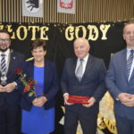 Na zdjęciu są cztery osoby w eleganckich strojach. Tło zdobią napisy Złote Gody oraz świąteczne dekoracje. Dominują kolory ciemne z elementami złota.