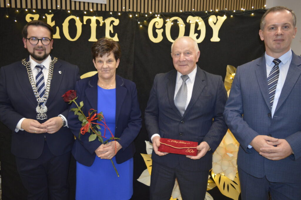 Na zdjęciu cztery osoby stoją na tle z napisem Złote Gody. Kobieta trzyma czerwoną różę, a mężczyzna etui. Wszyscy są ubrani elegancko.