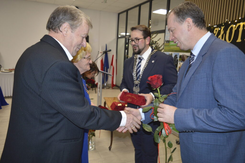Mężczyzna w garniturze wręcza różę i odznaczenie starszemu panu w sali z flagami. Tło jest jasne, z dekoracjami. Obok starsza kobieta patrzy z uśmiechem.