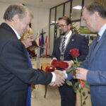 Mężczyzna w garniturze wręcza różę i odznaczenie starszemu panu w sali z flagami. Tło jest jasne, z dekoracjami. Obok starsza kobieta patrzy z uśmiechem.