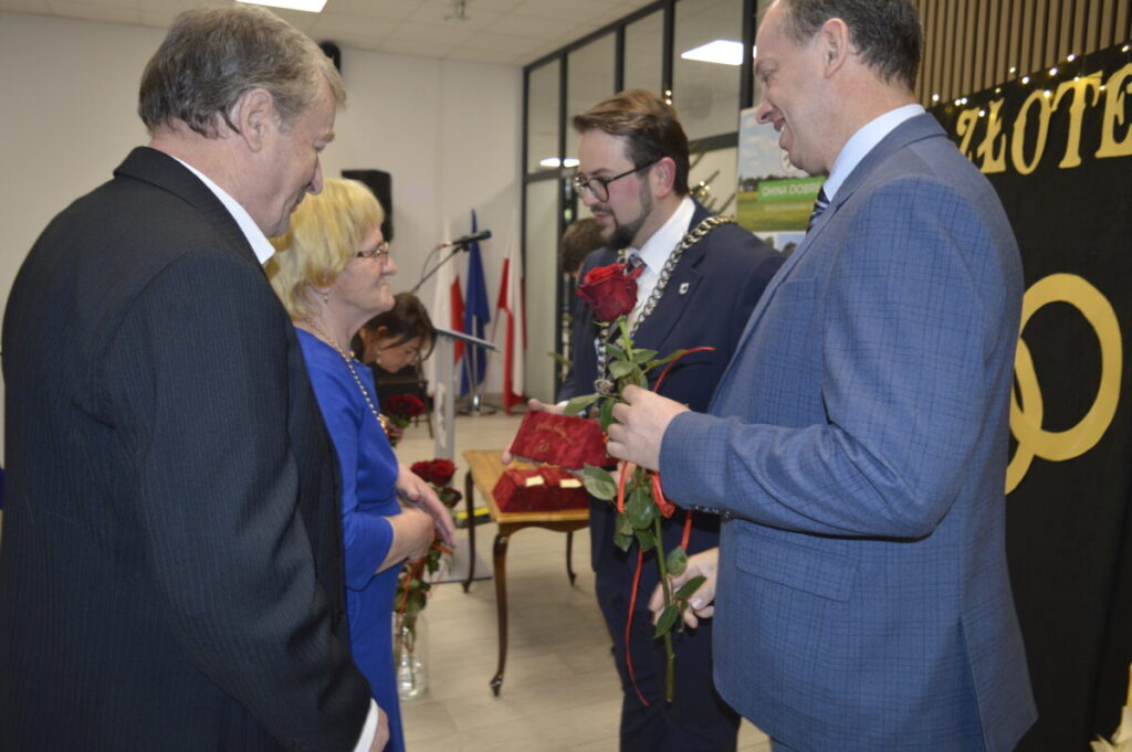 Na zdjęciu cztery osoby w formalnych strojach wymieniają się różami w jasnym pomieszczeniu. Tło zdobią flagi i czarny plakat z napisem. Atmosfera jest uroczysta.