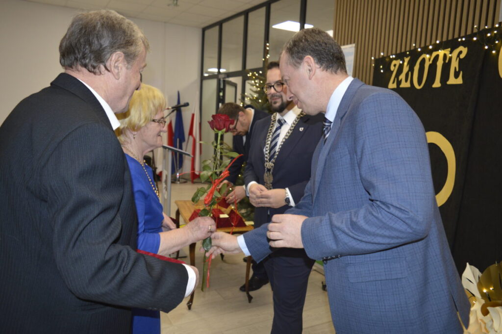 Grupa osób ubranych elegancko, mężczyzna wręcza kobiecie różę. Tło jasne, napisy „Złote” w tle. Widać flagi i mikrofony.