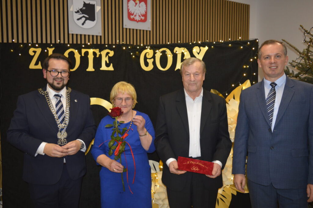 Cztery osoby stojące na tle czarnego baneru z napisem ZŁOTE GODY, z lampkami. Kobieta trzyma czerwoną różę.