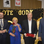 Cztery osoby stojące na tle czarnego baneru z napisem ZŁOTE GODY, z lampkami. Kobieta trzyma czerwoną różę.