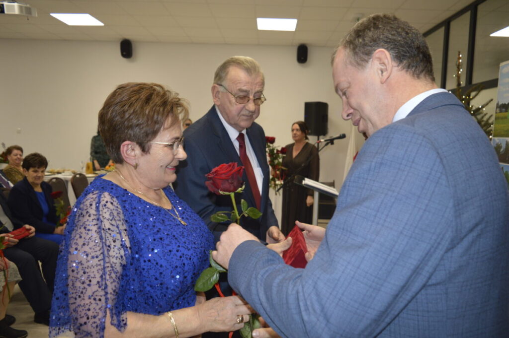 Starsza kobieta w niebieskiej sukience przyjmuje czerwoną różę od mężczyzny w szarym garniturze, obok stoi starszy mężczyzna.