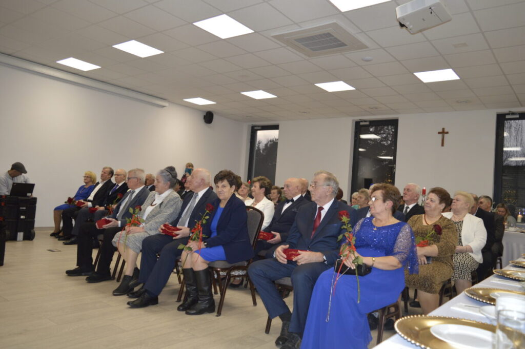 Grupa elegancko ubranych osób siedzi w sali z jasnym sufitem. Trzymają czerwone róże, tło to białe ściany z krzyżem. Obok stoły z zastawą.