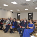 Grupa elegancko ubranych osób siedzi w sali z jasnym sufitem. Trzymają czerwone róże, tło to białe ściany z krzyżem. Obok stoły z zastawą.