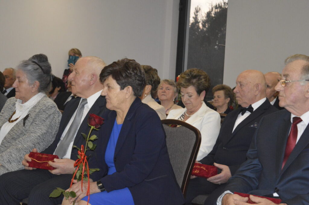 Grupa elegancko ubranych osób siedzi w sali, trzymając czerwone róże i kubki z napisami. Tło jest jasne i neutralne. Event w formalnym otoczeniu.