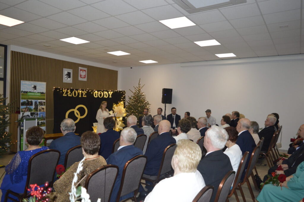 Sala z jasnym sufitem, ludzie siedzący, udekorowana choinka. Na tle tablica z napisem ZŁOTE GODY i symboliczne obrączki.