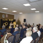 Sala z jasnym sufitem, ludzie siedzący, udekorowana choinka. Na tle tablica z napisem ZŁOTE GODY i symboliczne obrączki.