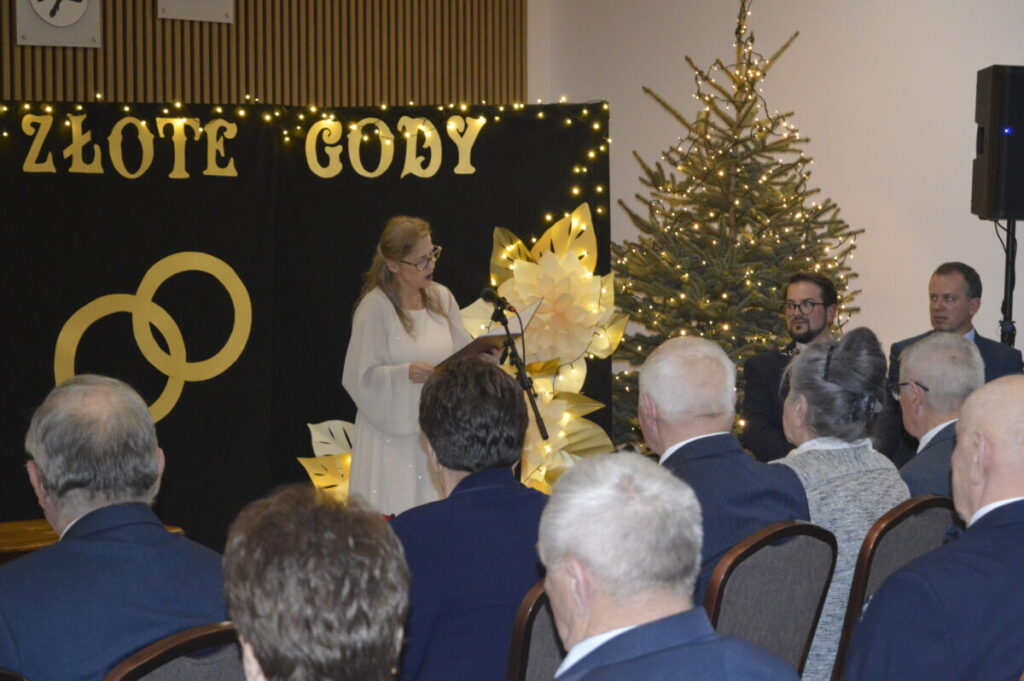 Kobieta w białej sukni przemawia na tle czarnego baneru z napisem ZŁOTE GODY. Obok niej stoi choinka ozdobiona światełkami. Wokół siedzą goście w eleganckich strojach.