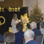 Kobieta w białej sukni przemawia na tle czarnego baneru z napisem ZŁOTE GODY. Obok niej stoi choinka ozdobiona światełkami. Wokół siedzą goście w eleganckich strojach.