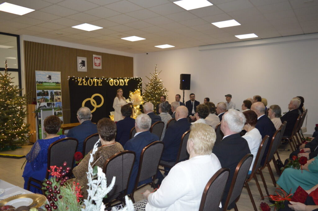 Sala z ludźmi na uroczystości Złote Gody. Występująca osoba na tle dekoracji z złotymi obrączkami. Świerki z lampkami.