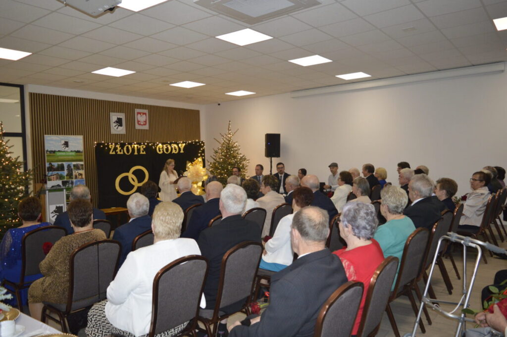 Ludzie siedzą na uroczystości Złote Gody. Tło to czarne zasłony z napisem, a obok stoją choinki. Jasne, przytulne wnętrze.