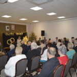 Ludzie siedzą na uroczystości Złote Gody. Tło to czarne zasłony z napisem, a obok stoją choinki. Jasne, przytulne wnętrze.