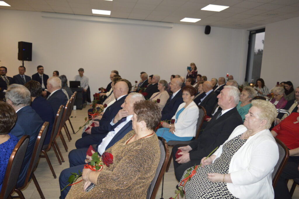 Grupa starszych osób siedzi w jasnej sali, trzymając czerwone róże. Ściany są białe, a krzesła brązowe. W tle widoczni stojący ludzie.