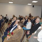 Grupa starszych osób siedzi w jasnej sali, trzymając czerwone róże. Ściany są białe, a krzesła brązowe. W tle widoczni stojący ludzie.