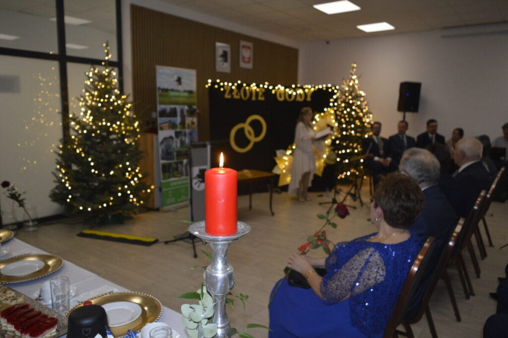 To zdjęcie z przyjęcia „Złote Gody”. Na pierwszym planie czerwono ubrana świeca, w tle choinki z lampkami. Goście siedzą przy stole, para obrączek na czarnym tle.
