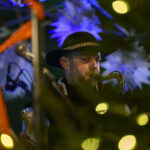 Muzyk w kapeluszu gra na saksofonie, otoczony świątecznymi dekoracjami. Dominują ciemne kolory z jasnymi akcentami. Tło jest rozmazane z żółtymi i niebieskimi światłami.