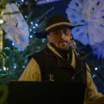 Mężczyzna w kapeluszu gra na gitarze, w tle zielona choinka z niebieskimi światełkami i papierowe ozdoby.