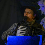 Mężczyzna w ciemnym kapeluszu gra na akordeonie, śpiewając do mikrofonu. Tło jest ciemne z białymi dekoracjami. Światło jest niebieskawe.