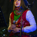 Osoba w kolorowym stroju ludowym trzyma mikrofon. Czerwony kamizelka, zielona chusta, ciemne tło. Sceniczne oświetlenie.