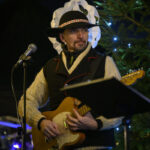 Mężczyzna w czarnym kapeluszu gra na gitarze w świątecznej scenerii. W tle zielone drzewko z lampkami. Jasne, ciepłe oświetlenie.