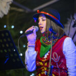 Kobieta w ludowym stroju śpiewa do mikrofonu. Ma na sobie kapelusz z czerwonym paskiem, na tle światełek. Obok stoi pulpit na nuty.