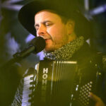 Mężczyzna gra na akordeonie marki Hohner i śpiewa do mikrofonu. Ma na sobie czarny kapelusz i szary sweter. Tło jest ciemne, z rozmytymi refleksami światła.