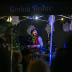 Na scenie kobieta w stroju ludowym stoi przed mikrofonem. Tło zdobią białe dekoracje i zielona choinka. Mroczny wieczór, napis nad sceną.