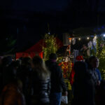 Nocny jarmark z oświetlonymi choinkami i tłumem ludzi. W tle czerwone stoiska i sceny ozdobione lampkami. Atmosfera świąteczna.