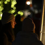 Dwie osoby w czapkach stoją przy grzejniku na zewnątrz. Ciepłe światło oświetla ich twarze, w tle rozmyte zielone światła.