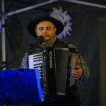 Muzyk gra na akordeonie marki Hohner, ubrany w ciemny kapelusz i sweter. Niebieskie oświetlenie i białe dekoracje w tle. Ciemne tło sceniczne.