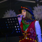 Osoba w ludowym stroju śpiewa do mikrofonu na scenie. Nosi czarno-czerwony kapelusz. Tło jest ciemne z białymi dekoracjami.