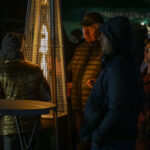 Ludzie w ciepłej odzieży gromadzą się wokół ogrzewacza na świeżym powietrzu nocą. Tło jest ciemne, co podkreśla ciepłe światło płomieni. Atmosfera zimowa, kameralna.