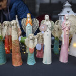 Rząd glinianych aniołów na czarnym tle stołu. Anioły w kolorach białym, turkusowym, różowym i pomarańczowym. W tle osoba i latarnia.