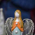 Anioł z jasnobrązowymi włosami ma pomarańczową suknię i białe, ażurowe skrzydła. Tło jest rozmyte, ciemnoniebieskie. Obok widoczna inna rzeźba.